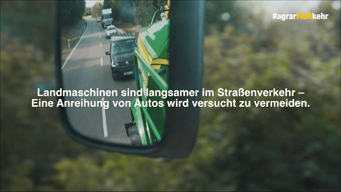 Ein Seitenspiegel zeigt Autos, die einem langsam fahrenden grünen landwirtschaftlichen Fahrzeug auf einer Landstraße folgen, mit deutscher Textüberlagerung.