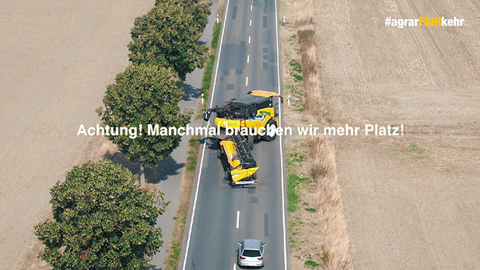 Ein breiter landwirtschaftlicher Wagen und ein Auto fahren auf einer Landstraße dicht aneinander vorbei, wobei ein Text darauf hinweist, dass mehr Platz benötigt wird.