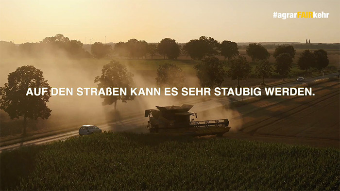 Ein Mähdrescher wirbelt Staub in der Nähe einer Straße mit Autos auf; ein deutscher Text überlagert die Szene bei Sonnenuntergang.