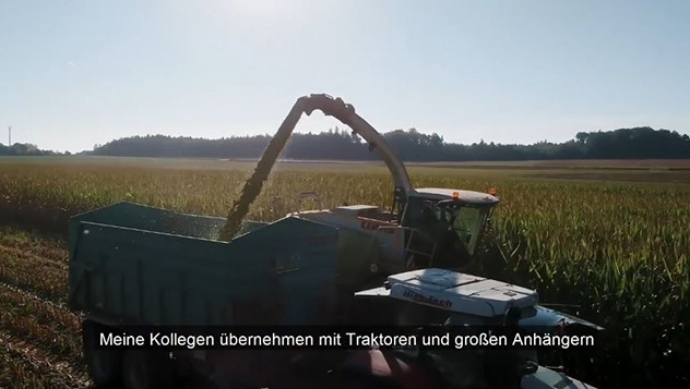 Ein Traktor erntet Mais auf einem Feld und füllt einen großen Anhänger; deutsche Untertitel werden angezeigt.