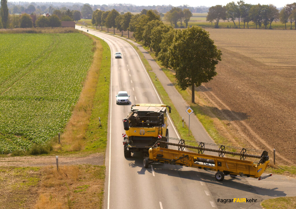 Ein großes landwirtschaftliches Fahrzeug blockiert eine Landstraße und zwingt die Autos zum Anhalten; auf beiden Seiten stehen Bäume.