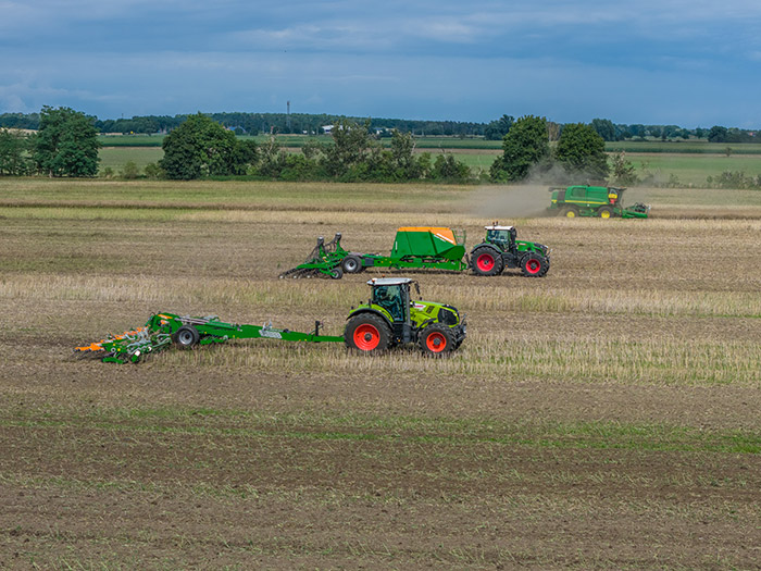 Drei Traktoren und ein Mähdrescher arbeiten auf einem großen, offenen Feld unter einem bewölkten Himmel.