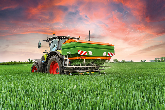 Ein Traktor düngt ein üppiges grünes Feld unter einem dramatischen rot-orangenen Sonnenuntergangshimmel.