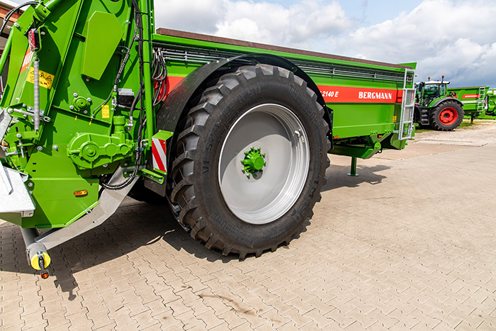 Grüner landwirtschaftlicher Anhänger mit großen schwarzen Reifen, geparkt auf einer gepflasterten Fläche unter einem bewölkten Himmel.