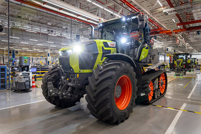 Ein großer grün-schwarzer Traktor mit eingeschalteten Scheinwerfern steht in einer hell erleuchteten Fabrik.