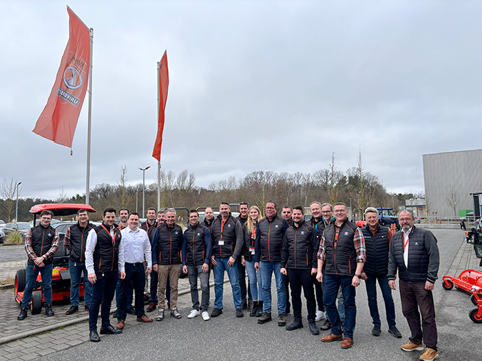 Eine Gruppe von Menschen posiert an einem bewölkten Tag im Freien neben roten Fahnen und Maschinen.