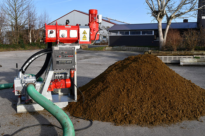 Rote Gülleseparatormaschine mit Schläuchen neben einem großen Güllehaufen auf einer Betonfläche im Freien.