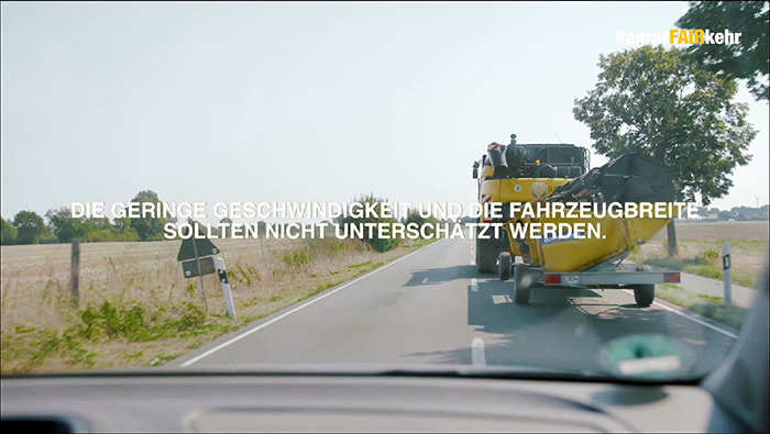 Ein Auto folgt einem breiten landwirtschaftlichen Fahrzeug auf einer Landstraße, wobei ein deutscher Text über Geschwindigkeit und Breite eingeblendet wird.