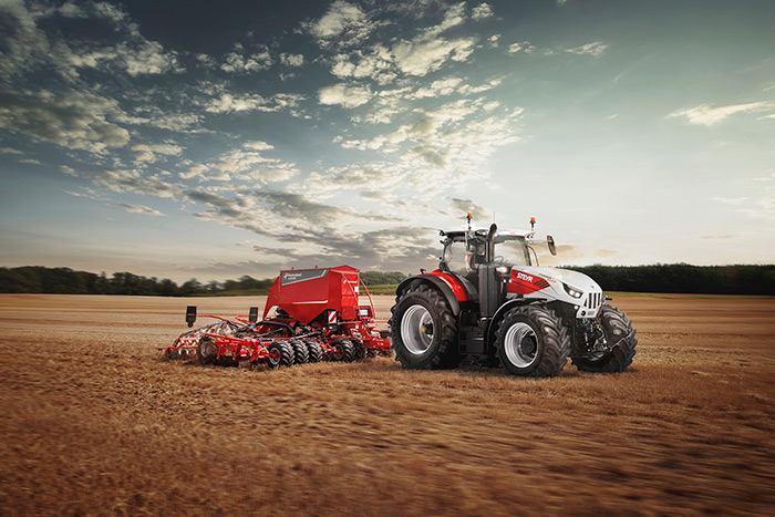 Ein rot-weißer Traktor, der unter einem teilweise bewölkten Himmel landwirtschaftliche Geräte über ein Feld zieht.