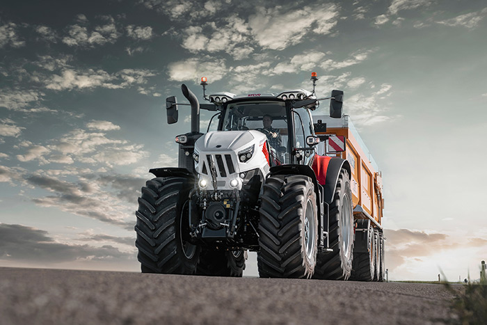 Ein moderner Traktor mit großen Reifen und einem Anhänger fährt auf einer Landstraße unter einem bewölkten Himmel.