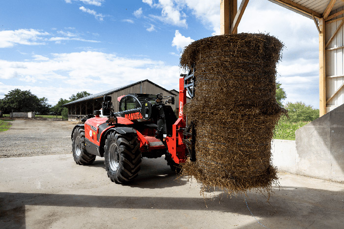Ein Traktor hebt an einem sonnigen Tag einen großen Rundballen Heu in einer Scheune.