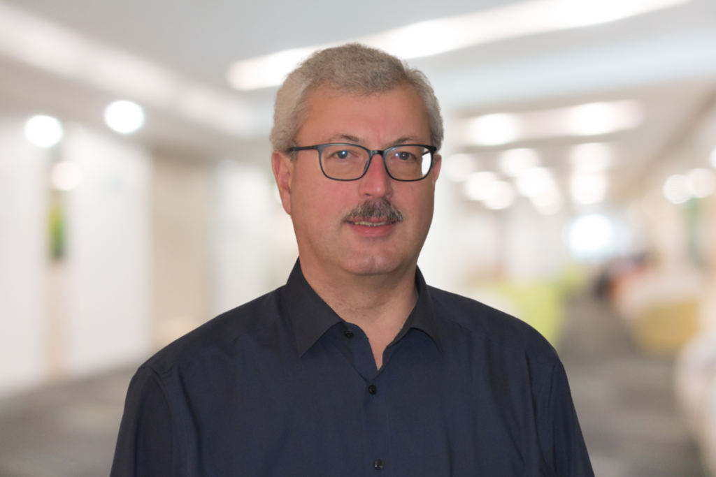 Ein Mann mit Brille und Schnurrbart, der ein dunkles Hemd trägt, steht in einem hell erleuchteten Korridor.