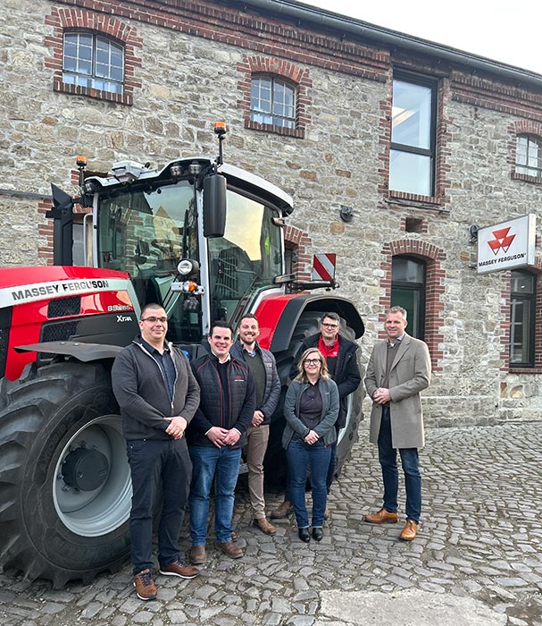 Sechs Personen stehen vor einem roten Traktor und einem Steingebäude mit Massey Ferguson-Schildern.